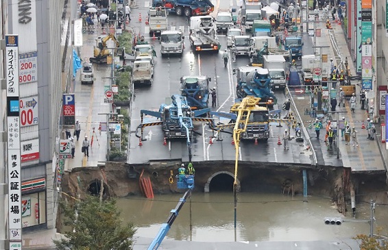 A giant sinkhole in a five-lane urban boulevard, eroding soil exposing underground steel columns supporting commercial buildings in Fukuoka, Southwest Japan