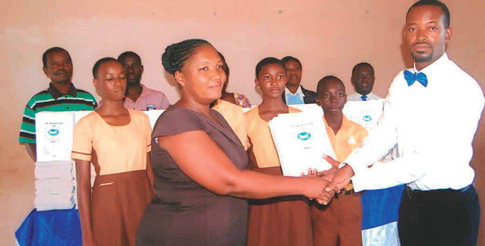  Mr Robert Mensah Aheto presenting some of the branded exercise books to Ms Emmanuella Tsyawo, with some pupils of the school looking on