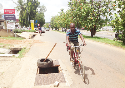 A cyclist riding past an uncovered underground utility vault of Vodafone Ghana along the Olusegun Highway at Pig Farm Junction