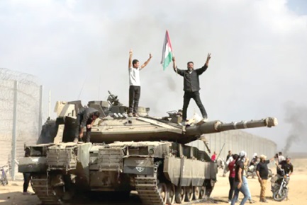 Palestinians wave their national flag and celebrate by a destroyed Israeli tank at the Gaza Strip fence east of Khan Younis on October 7, 2023 (Credit: Yousef Masoud, AP Photo, Aljazeera)