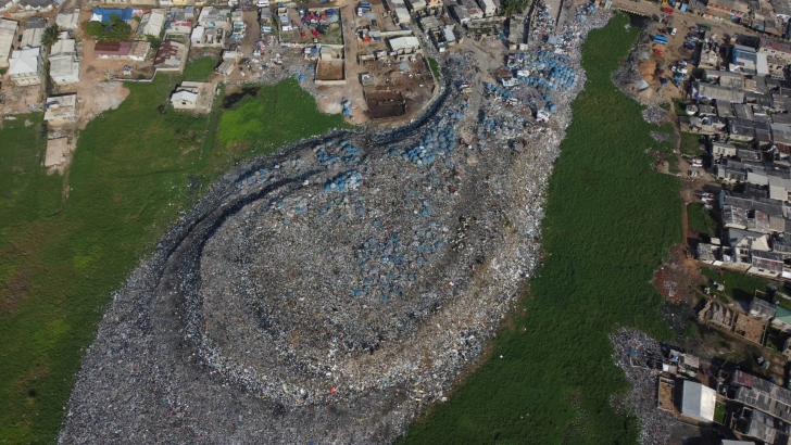 An aerial photograph of a roughly 4-year-old textile dump site in Glefe, inside the Densu Delta protected wetlands, outside Accra, Ghana. The dump site sits close to lagoons and streams, with no lining and no other visible pollution mitigation systems.