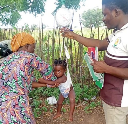 A health worker weighing a baby under a neem tree.