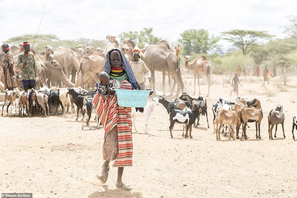 A young girl carries a baby and a tub of water away from the borehole as goats and camels take their turn to drink
